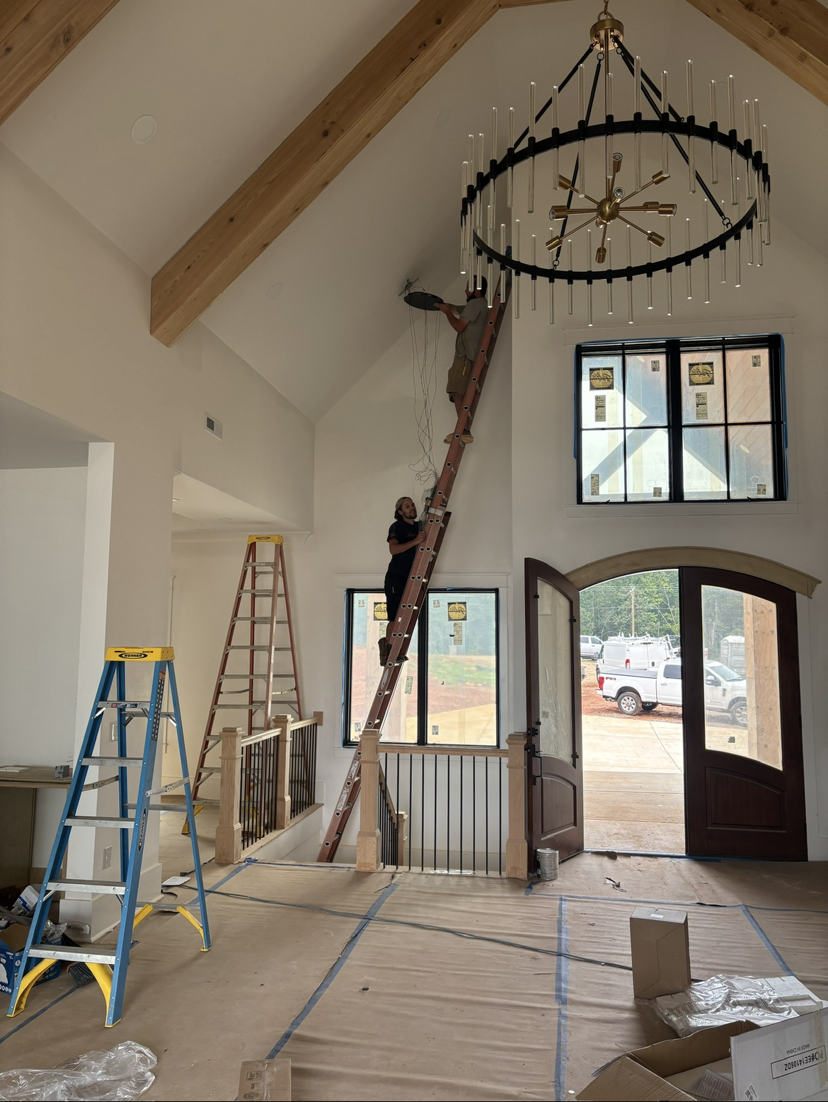 A person stands on a tall ladder working near the ceiling of a large, unfinished room with exposed wooden beams and a large chandelier. Tools, ladders, and boxes are visible.