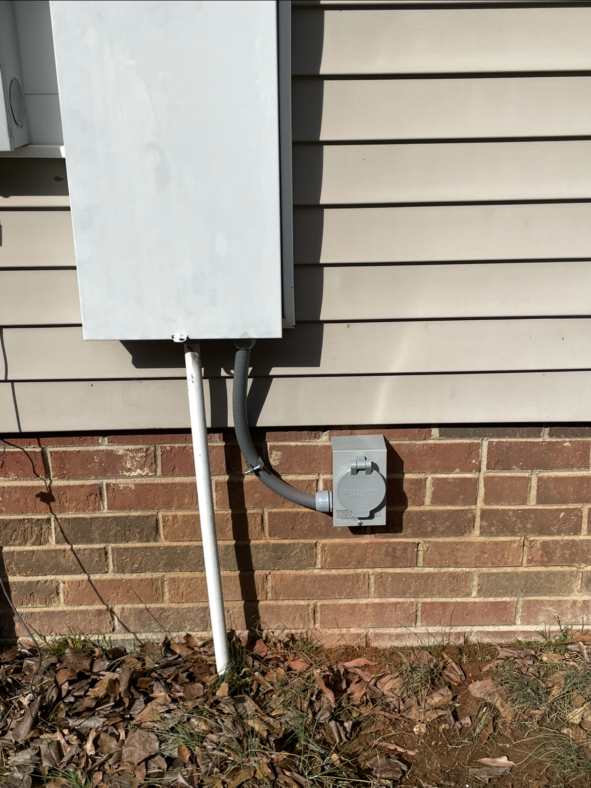 A gray electrical panel and a smaller outlet box, ideal for generator services, are mounted on the exterior wall of a house with beige siding and a brick foundation.