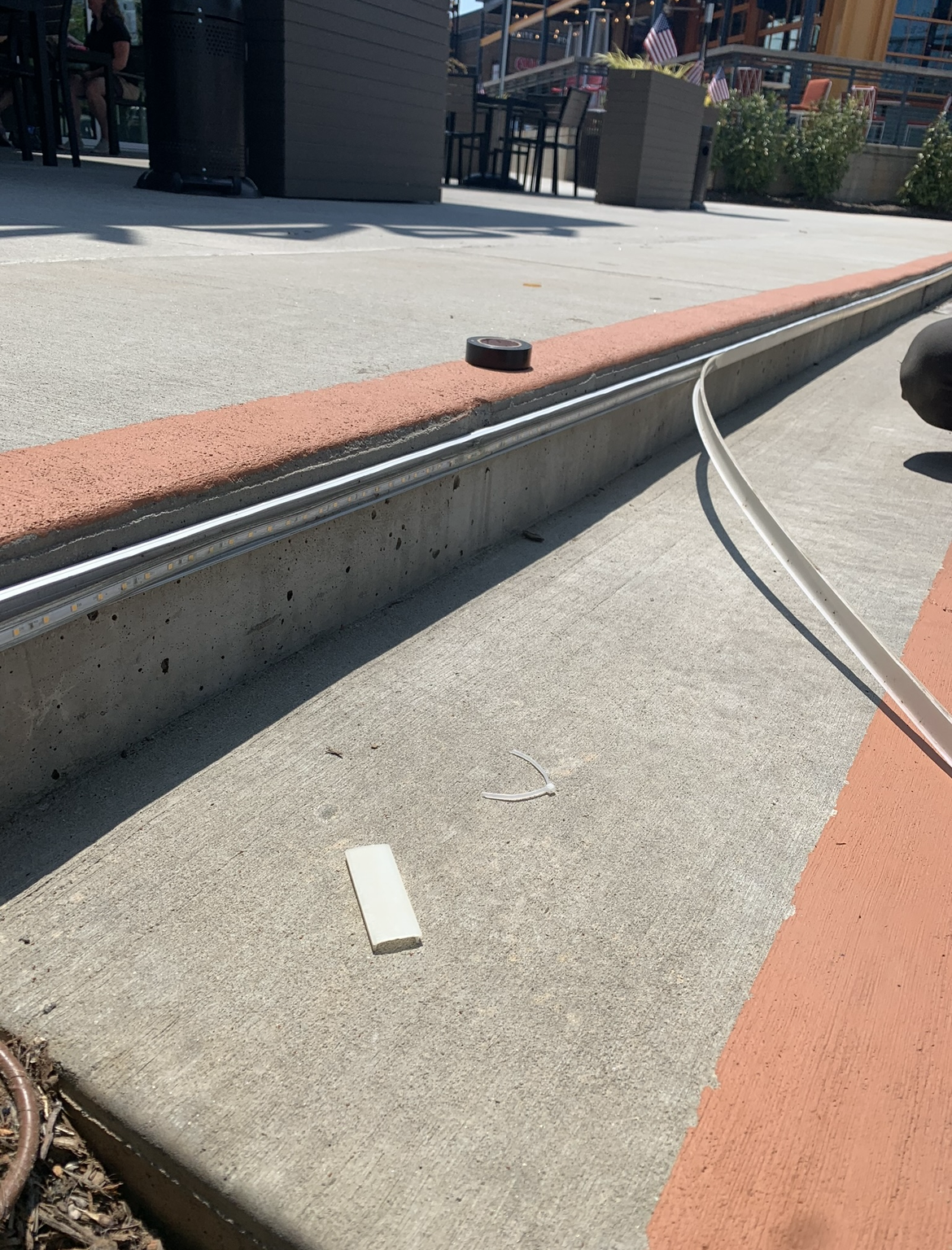 A concrete sidewalk with an orange border, a metal rail, a white rectangular object possibly related to generator services, a cable, and outdoor seating in the background.
