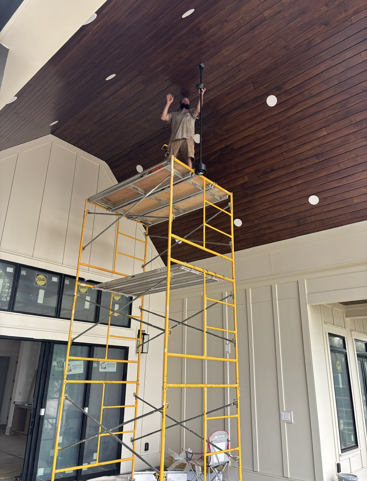 A person stands on yellow scaffolding, using a tool to work on a dark wood ceiling in a partially finished room with cream walls and large windows, possibly preparing the space for electrical installations or generator services.