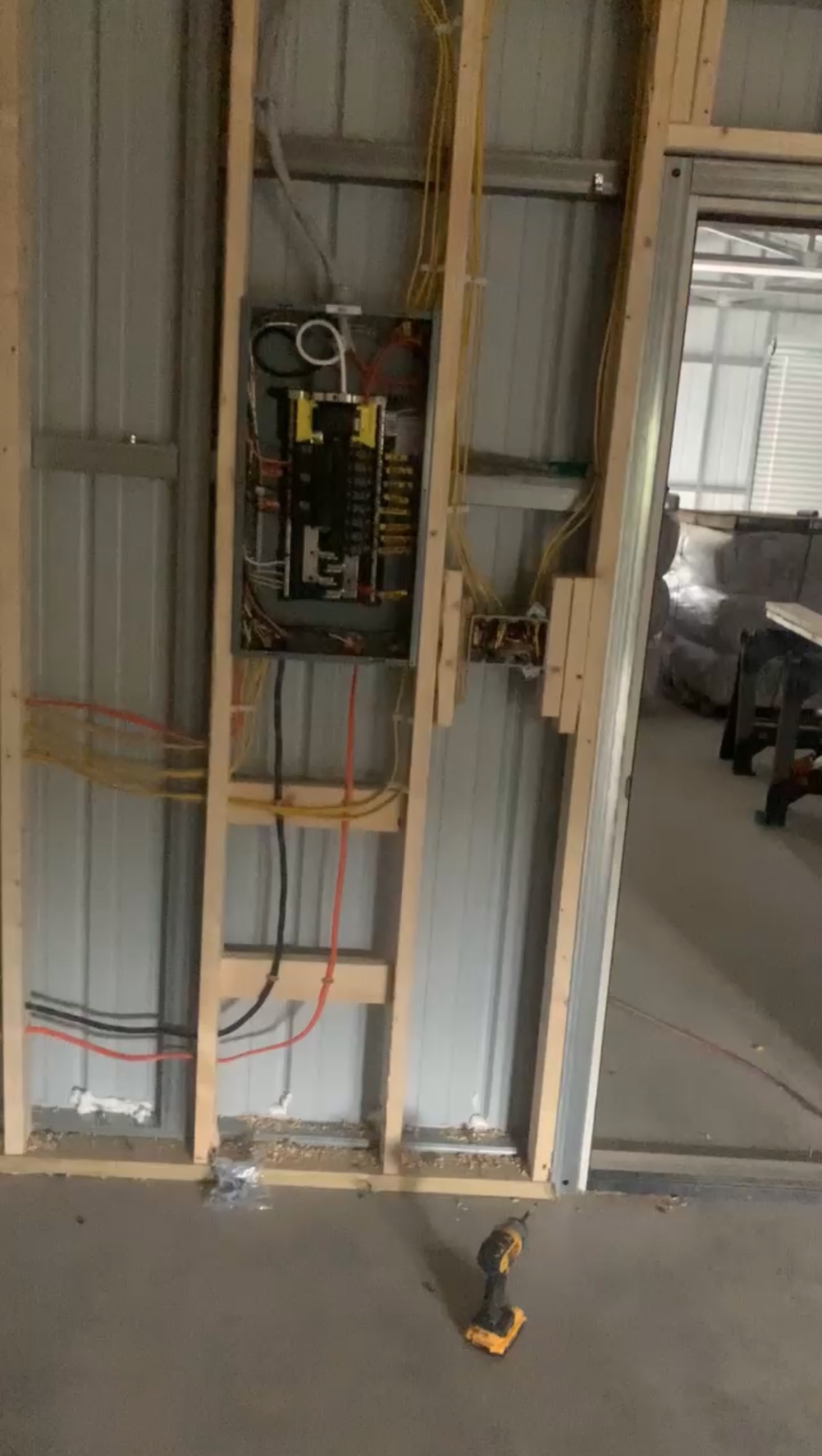 Electrical panel with exposed wiring mounted on a metal wall frame next to a doorway in a partially constructed room—ideal for integrating generator services. A power drill is on the floor nearby.