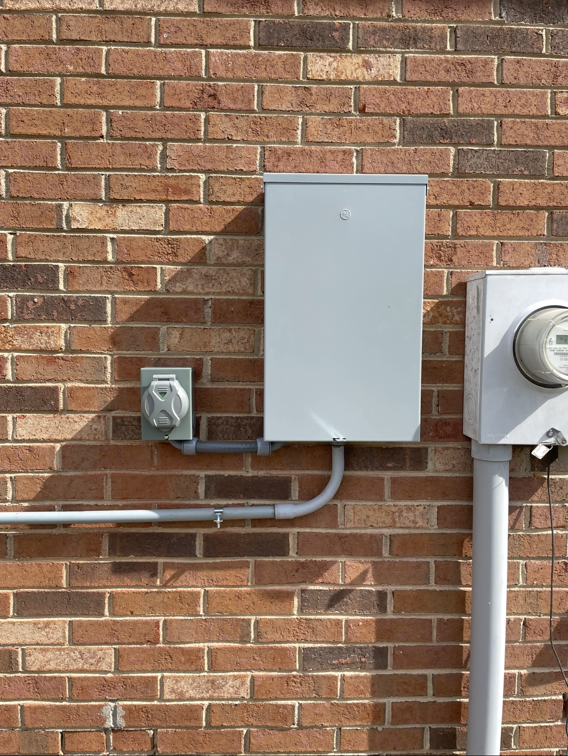 Electrical box, outlet, and meter mounted on a red brick wall, with metal conduit pipes running along the surface—ideal for integrating generator services to ensure reliable backup power.